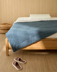 Blue quilt blanket with a textured surface on a wooden bed frame, with a white pillow and beige wooden wall background