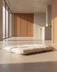 A white quilted mattress topper or mattress pad with a thin gold trim, shown resting on a light gray floor in front of a wooden cabinet.